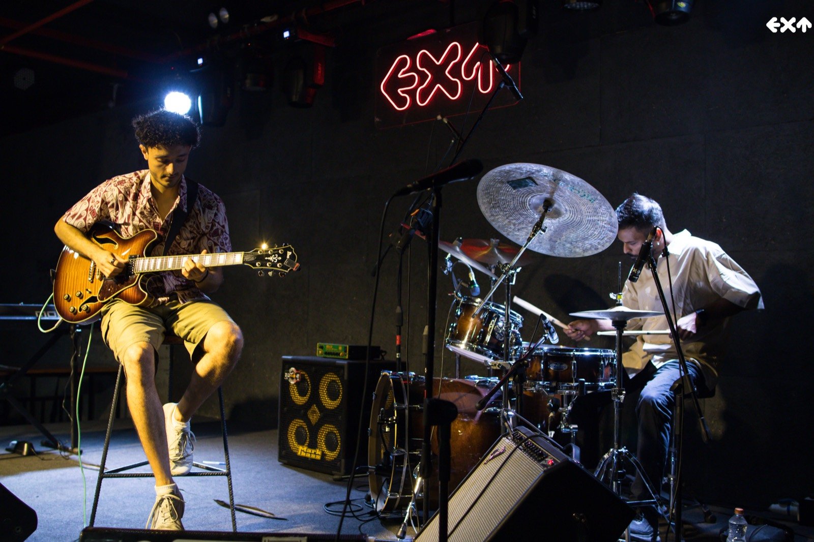 Lucas Baret seated on a club stage playing the archtop, a drummer beside him under a neon sign